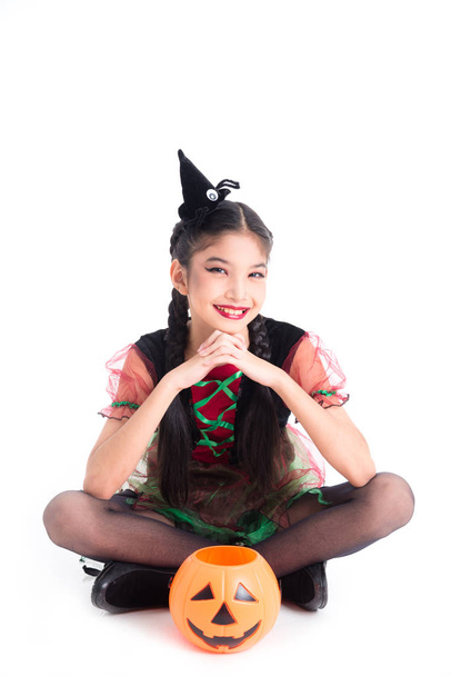 Pełna długość małej azjatyckiej dziewczyny w stroju czarownicy siedzi i uśmiecha się z Halloweenowym wiadrem dyni - Zdjęcie, obraz