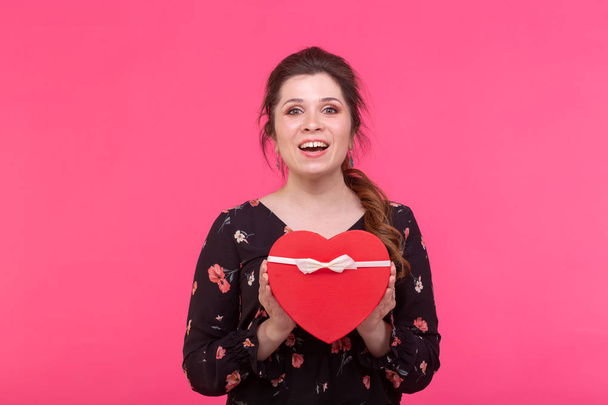 Holidays, present and surprise concept - Beauty happy girl with Valentine Gift box on pink background. Smiling surprised model girl takes heart shaped red present. - Photo, Image