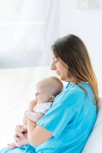 πλευρική όψη της ελκυστικής και νεαρής μητέρας που κρατά το παιδί της στο νοσοκομείο  - Φωτογραφία, εικόνα