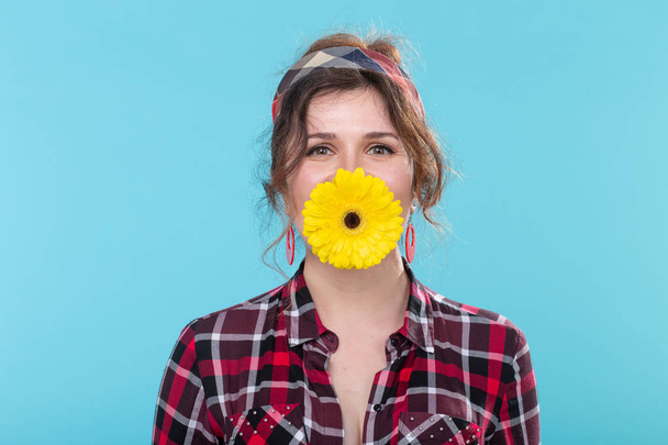 Funny positive young woman in retro image holding a bright yellow gerbera flower in her teeth posing against a blue background. Concept of surprise and fun. - Foto, Imagem