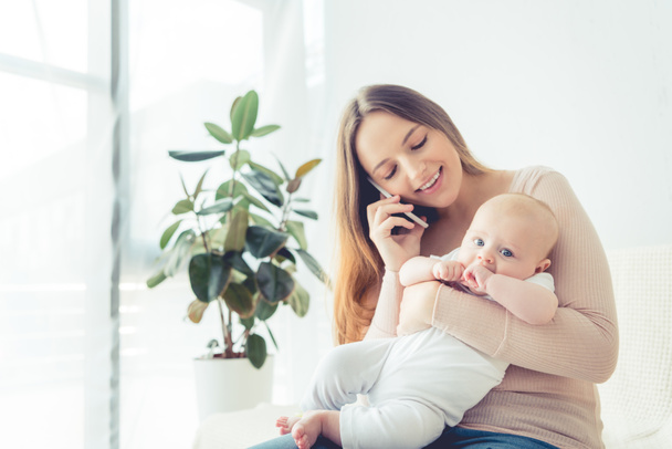 ελκυστική και χαμογελαστή μητέρα κρατώντας το παιδί της και μιλώντας στο smartphone στο διαμέρισμα  - Φωτογραφία, εικόνα