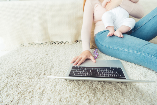 oříznutý pohled matky držící dítě a používání laptopu v bytě  - Fotografie, Obrázek
