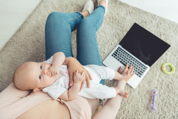 Ausgeschnittene Ansicht einer Mutter, die ihr Kind hält und Laptop in Wohnung benutzt  - Foto, Bild