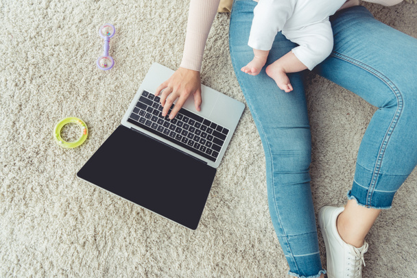περικομμένη θέα της μητέρας κρατώντας το παιδί της και τη χρήση του φορητού υπολογιστή στο διαμέρισμα  - Φωτογραφία, εικόνα