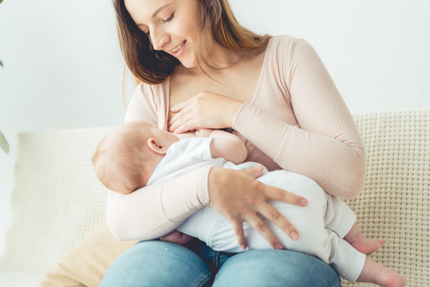 attractive and smiling mother breastfeeding her child in apartment  - Фото, зображення