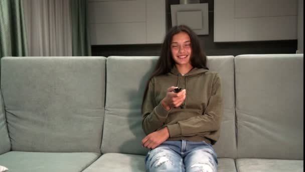 pretty young lady in hoodie and jeans watches television - Materiał filmowy, wideo