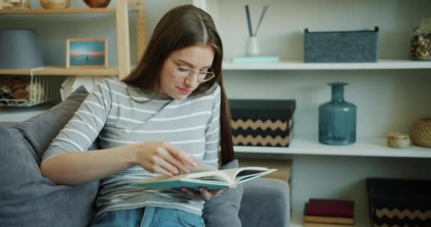 Slow motion of pretty young lady smiling reading book on sofa at home - Video
