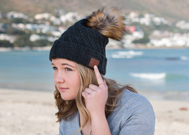 Modelo femenino con gorro de punto gris con altavoces Bluetooth en el interior, escuchando música al aire libre. Con el dedo en los botones
. - Foto, Imagen
