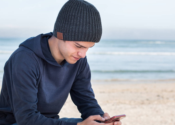 Modelo masculino vestindo gorro de malha cinza escuro com alto-falantes Bluetooth dentro, ouvindo música com telefone inteligente móvel ao ar livre. Olhando para o dispositivo móvel
. - Foto, Imagem