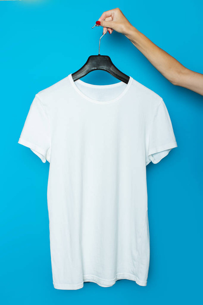 Vrouwelijke hand houdt een wit T-shirt zonder een foto op een trempel op een blauwe achtergrond - Foto, afbeelding