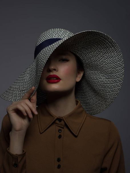 Beautiful woman portrait with red lipstick on lips and a big hat. Vintage image of a mysterious girl. Fashionable makeup. The hat covers half the face. Black eyeliner and extremely long eyelashes - 写真・画像