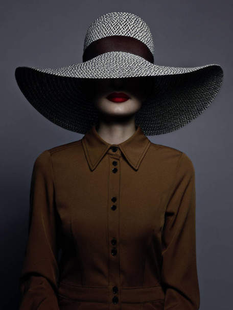 Beautiful woman portrait with red lipstick on lips and a big hat. Vintage image of a mysterious girl. Fashionable makeup and clothes. The hat covers half the face. Gray background - Fotó, kép