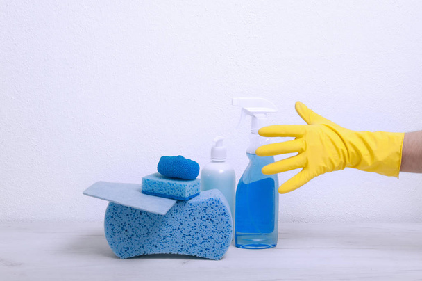 Gąbki, szale, detergenty na szarym stole na tle białej ścianie. Facet w żółtej rękawicy bierze spray do czyszczenia. - Zdjęcie, obraz