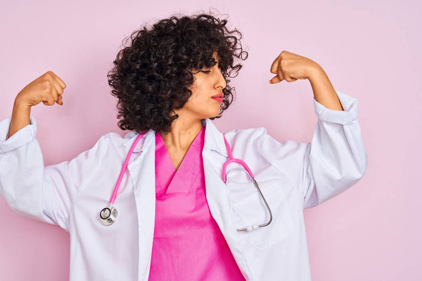 孤立したピンクの背景の上に聴診器を身に着けている巻き毛を持つ若いアラブの医師の女性は、腕の筋肉が誇らしげに微笑んでいます。フィットネスコンセプト. - 写真・画像
