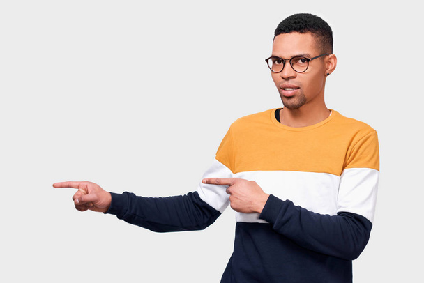 Οριζόντια πορτρέτο του έξυπνου Αφρικανού αμερικανικού νεαρού άνδρα δείχνει με τους δείκτη σε κενό χώρο αντίγραφο, φοράει πολύχρωμο πουλόβερ και γυαλιά, έχει σοβαρή έκφραση. Διαφήμιση και εθνικότητα  - Φωτογραφία, εικόνα