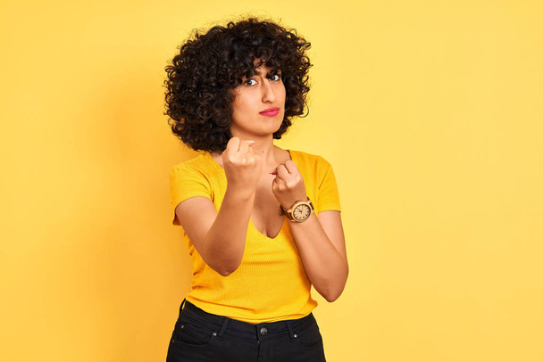 Jonge Arabische vrouw met krullend haar dragen t-shirt staande over geïsoleerde gele achtergrond klaar om te vechten met vuist verdediging gebaar, boos en boos gezicht, bang voor probleem - Foto, afbeelding