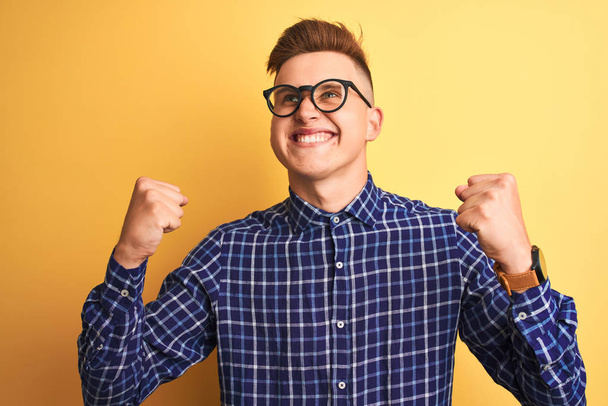 Young handsome man wearing casual shirt and glasses over isolated yellow background very happy and excited doing winner gesture with arms raised, smiling and screaming for success. Celebration concept. - Foto, imagen