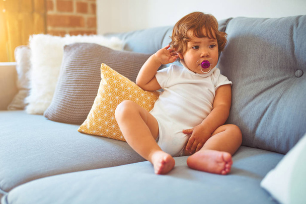 Красивая девочка-младенец в белой футболке сидит на диване, используя пустышку
 - Фото, изображение