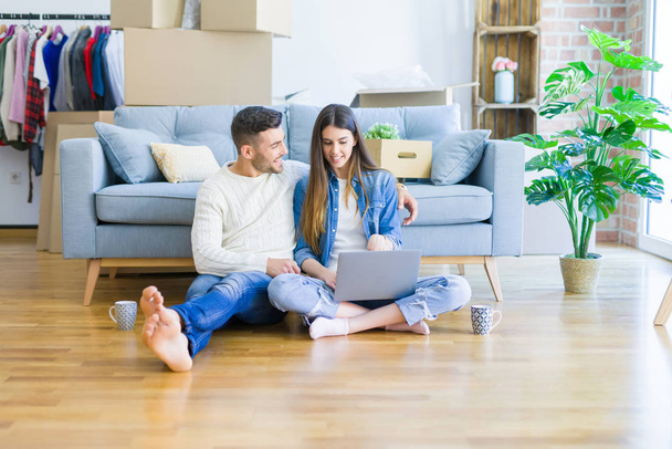 Jovem casal sentado no chão da nova casa usando laptop de computador e sorrindo feliz por se mudar para uma nova casa
 - Foto, Imagem