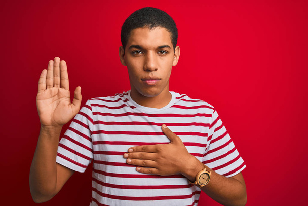 Young handsome arab man wearing striped t-shirt over isolated red background Swearing with hand on chest and open palm, making a loyalty promise oath - Photo, Image