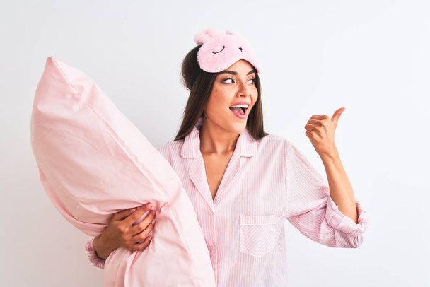 Mulher bonita usando máscara de sono pijama segurando travesseiro sobre fundo branco isolado apontando e mostrando com o polegar até o lado com o rosto feliz sorrindo
 - Foto, Imagem