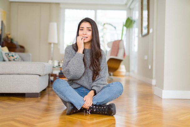 Mujer hermosa joven sentada en el suelo en casa mirando estresada y nerviosa con las manos en la boca mordiendo las uñas. Problema de ansiedad
. - Foto, imagen