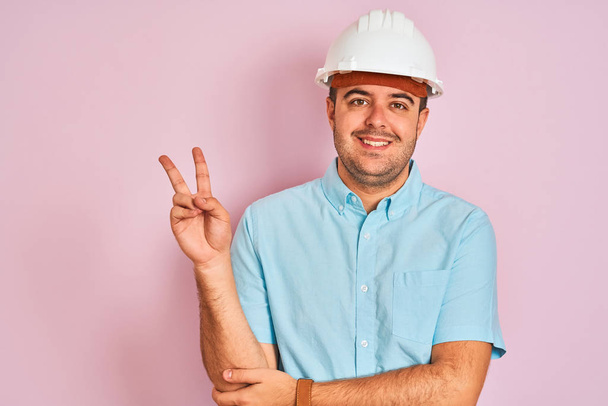 Young architect man wearing security helmet standing over isolated pink background smiling with happy face winking at the camera doing victory sign. Number two. - Φωτογραφία, εικόνα