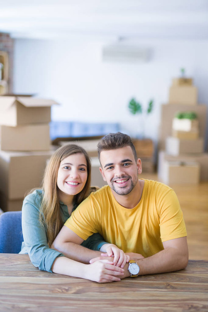 Young beautiful couple sitting on the table at home, hugging in love very happy for moving to new home with cardboard boxes behind them - Фото, зображення