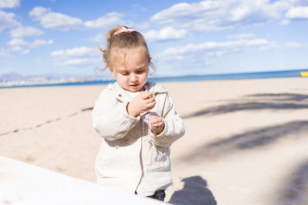 Kaunis lapsi lapsi tyttö yllään takki leikkii hiekalla rannalla
 - Valokuva, kuva