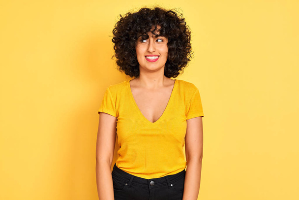 Nuori arabi nainen kihara tukka yllään t-paita seisoo yli eristetty keltainen tausta hymyillen katsoen sivuun ja tuijottaen pois ajattelu
. - Valokuva, kuva