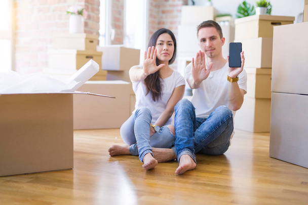 Όμορφο ζευγάρι κάθεται στο πάτωμα κρατώντας smartphone σε νέο σπίτι γύρω από χαρτόνι κουτιά με ανοιχτό χέρι κάνει στοπ σήμα με σοβαρή και αυτοπεποίθηση έκφραση, άμυνα χειρονομία - Φωτογραφία, εικόνα