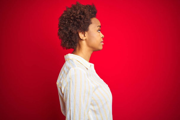 孤立した赤い背景の上にアフロヘアを持つ若い美しいアフリカ系アメリカ人女性が横に見て、自信のある笑顔で自然な顔でプロフィールポーズをリラックス. - 写真・画像