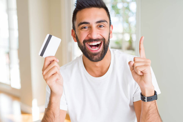 Jóképű hispán ember Holding hitelkártya meglepett egy ötlet vagy kérdés mutatva ujját a boldog arc, számú - Fotó, kép