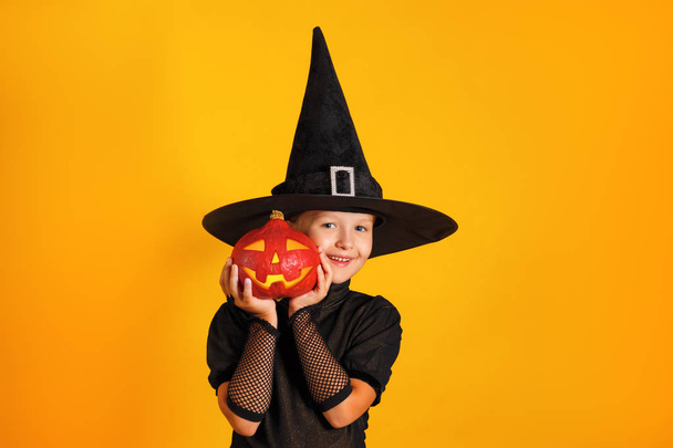Cute little girl dressed in a witch costume holds a pumpkin jack lantern on a yellow background. Halloween celebration - Foto, Imagen