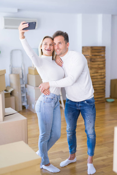 Young beautiful couple standing using smartphone to take selfie at new home around cardboard boxes - 写真・画像