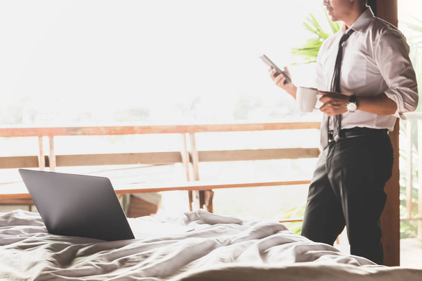 Фокус ноутбук на кровати с бизнесменом с помощью мобильного телефона и проведение кофе в чашке
. - Фото, изображение