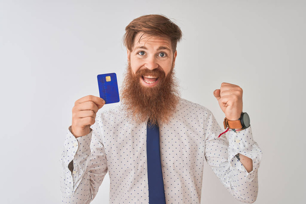 junger rothaariger irischer Geschäftsmann mit Kreditkarte vor isoliertem weißem Hintergrund, der stolz schreit und Sieg und Erfolg feiert, sehr aufgeregt, jubelnde Emotion - Foto, Bild