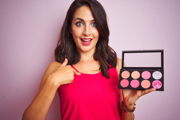 Jonge mooie vrouw aanbrengen make-up over roze geïsoleerde achtergrond met verrassing gezicht wijzende vinger naar zichzelf - Foto, afbeelding