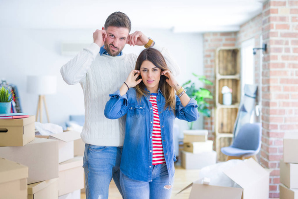 Young beautiful couple standing at new home around cardboard boxes covering ears with fingers with annoyed expression for the noise of loud music. Deaf concept. - Photo, Image