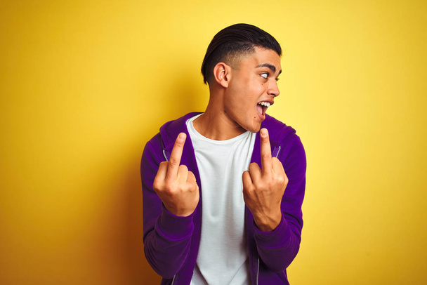 Jeune homme brésilien portant sweat-shirt violet debout sur fond jaune isolé montrant majeur faisant baise vous mauvaise expression, provocation et attitude grossière. Cris excités
 - Photo, image