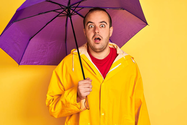 Νέος άντρας φορώντας αδιάβροχο κρατώντας μωβ ομπρέλα στέκεται πάνω από απομονωμένο κίτρινο φόντο φοβισμένος σε σοκ με μια έκπληξη πρόσωπο, φοβισμένος και ενθουσιασμένος με την έκφραση του φόβου - Φωτογραφία, εικόνα