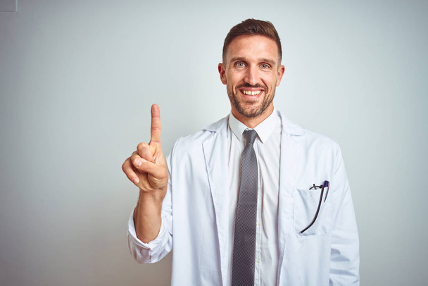Fiatal csinos orvos férfi visel fehér profressional kabát alatt elszigetelt háttér és a mutató felfelé ujjal számú miközben mosolyogva magabiztos és boldog. - Fotó, kép