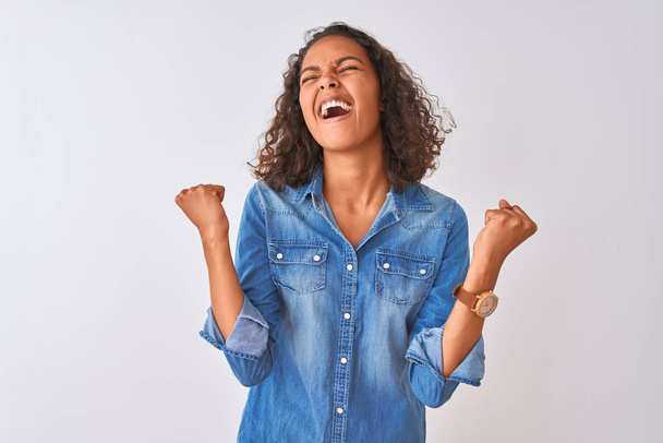 Young brazilian woman wearing denim shirt standing over isolated white background celebrating surprised and amazed for success with arms raised and eyes closed. Winner concept. - Photo, Image