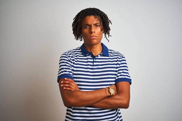 Afro man with dreadlocks wearing striped blue polo standing over isolated white background skeptic and nervous, disapproving expression on face with crossed arms. Negative person. - Photo, Image