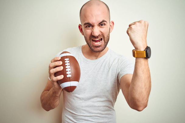 Mladý muž, který drží Rugby amerického fotbalového míče přes izolované pozadí, rozčilené a frustrovaný pokřik, šílený a křičící s zvedanou rukou, koncept hněvu - Fotografie, Obrázek