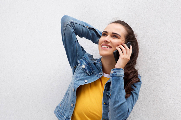 Retrato de una hermosa joven con el pelo castaño sonriendo y hablando con el teléfono móvil por la pared blanca
 - Foto, imagen