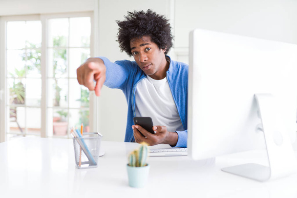 African American Man pracy przy użyciu smartfona i komputera wskazując palcem do aparatu i do Ciebie, ręka znak, pozytywny i pewny gest z przodu - Zdjęcie, obraz