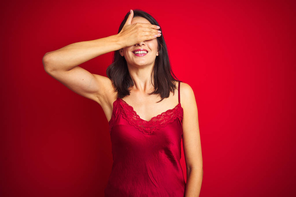 Молода красива жінка в сексуальній білизні над червоним ізольованим тлом посміхається і сміється з рукою на обличчі, прикриваючи очі сюрпризом. Сліпа концепція
. - Фото, зображення