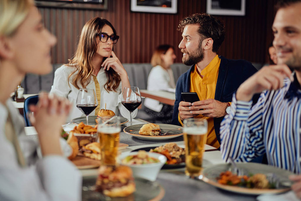 Amigos multiétnicos sentados en el restaurante, bebiendo alcohol, charlando y comiendo hamburguesas para cenar
. - Foto, Imagen