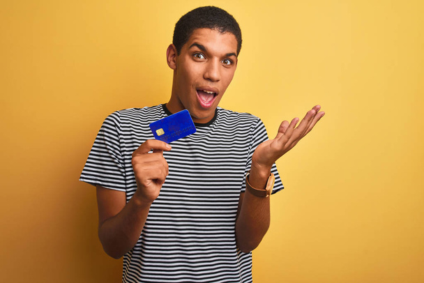 Joven hombre árabe guapo sosteniendo la tarjeta de crédito de pie sobre el fondo amarillo aislado muy feliz y emocionado, la expresión ganadora celebrando la victoria gritando con una gran sonrisa y las manos levantadas
 - Foto, imagen